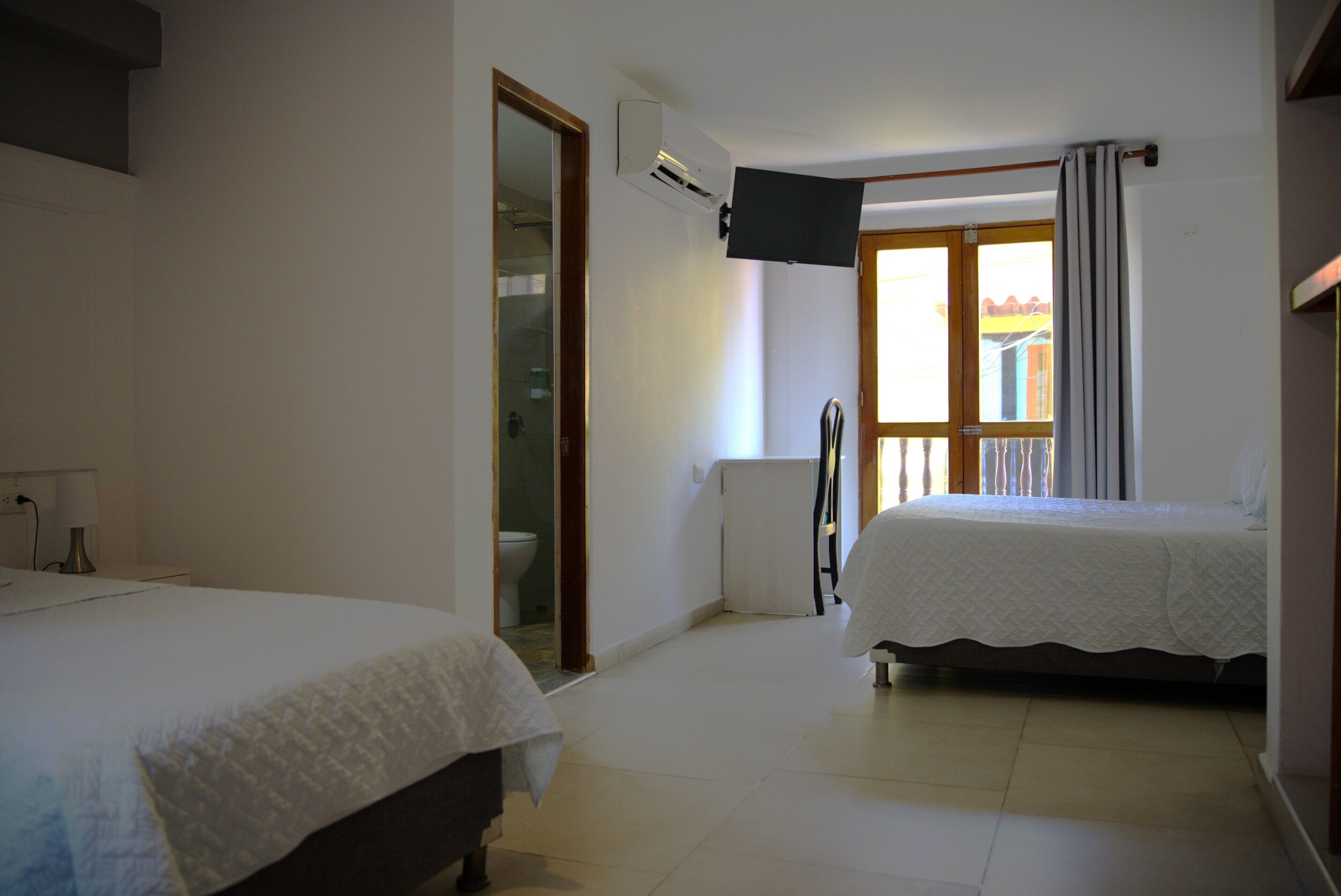 A hotel room with two beds, a TV mounted on the wall, a window with curtains, and a door leading to a bathroom.