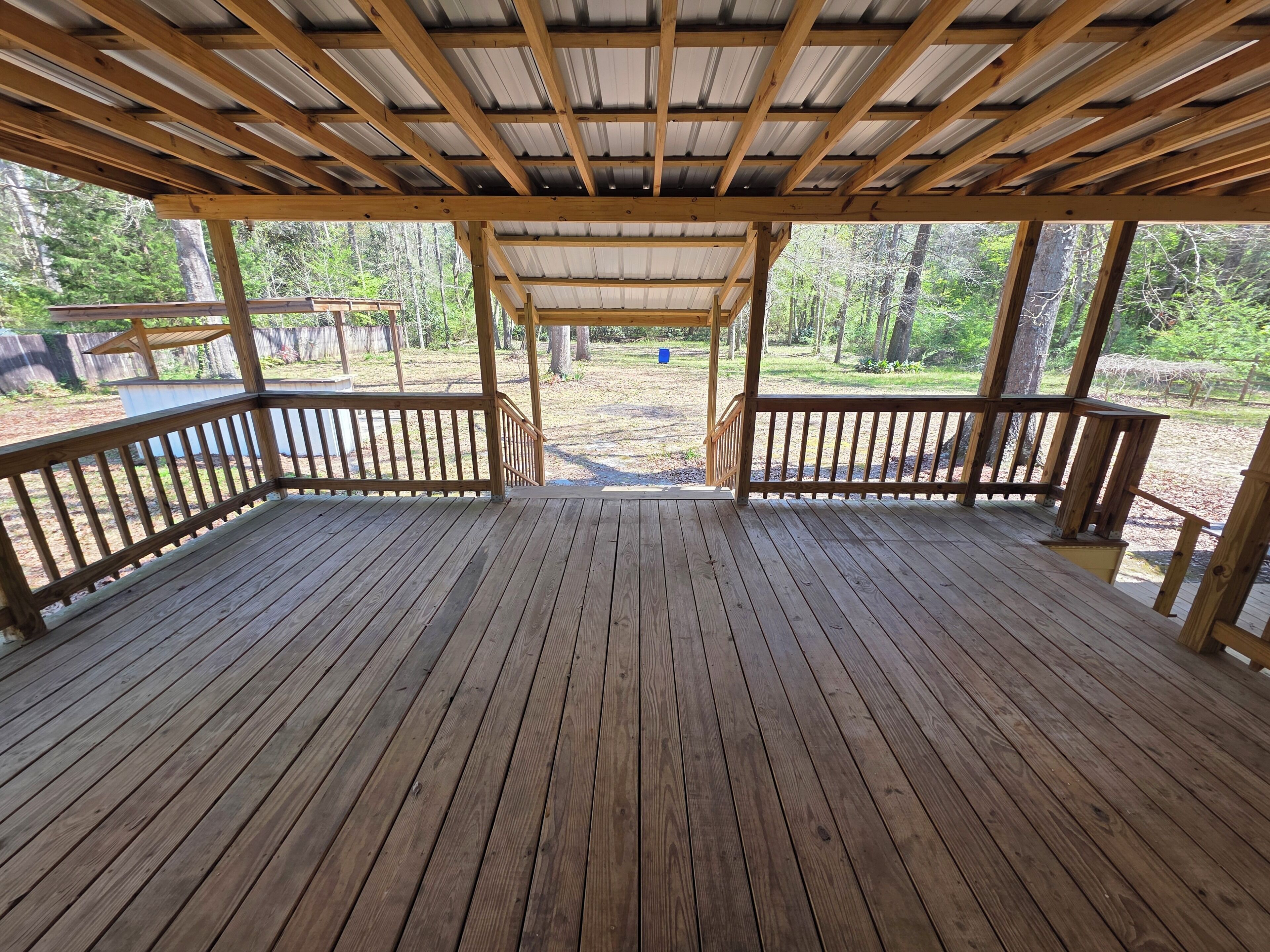 A wooden deck with a metal roof, surrounded by a wooden railing, and a view of a forested area.