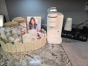 A kitchen counter with a coffee maker, mugs, a cookbook, and a basket of tea bags. - Padgett Peaceful Acres (Andalusia)