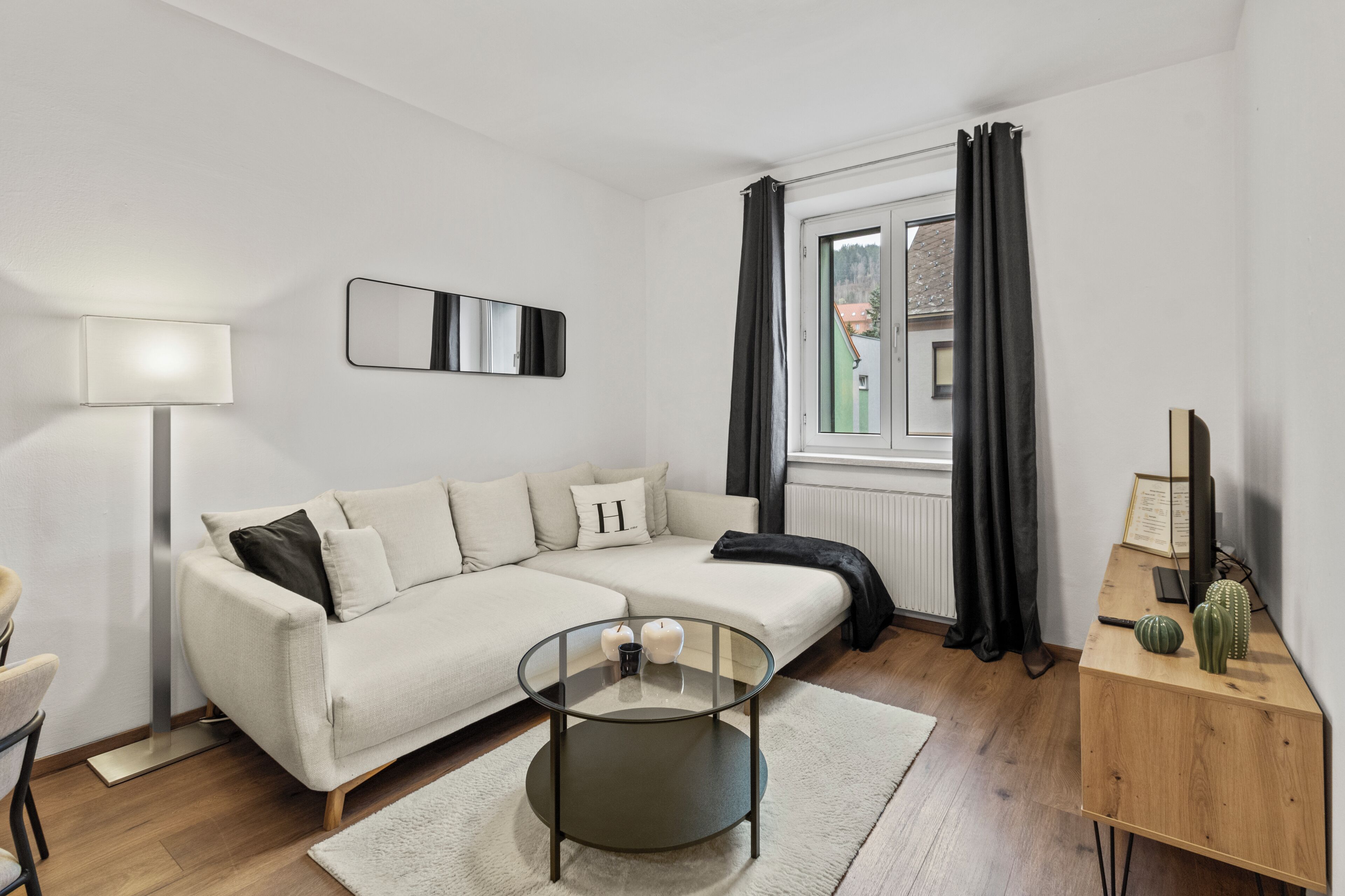 A modern living room with a sofa, a glass coffee table, a lamp, a wooden side table, and a window with curtains.