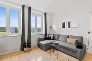 A modern living room with a grey sofa, a small side table, a large window with curtains, and a view of the outdoors.