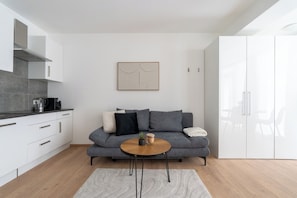 Un espacio habitable moderno con un sofá gris, una mesa de centro redonda de madera y una zona de cocina con armarios blancos y encimera gris.