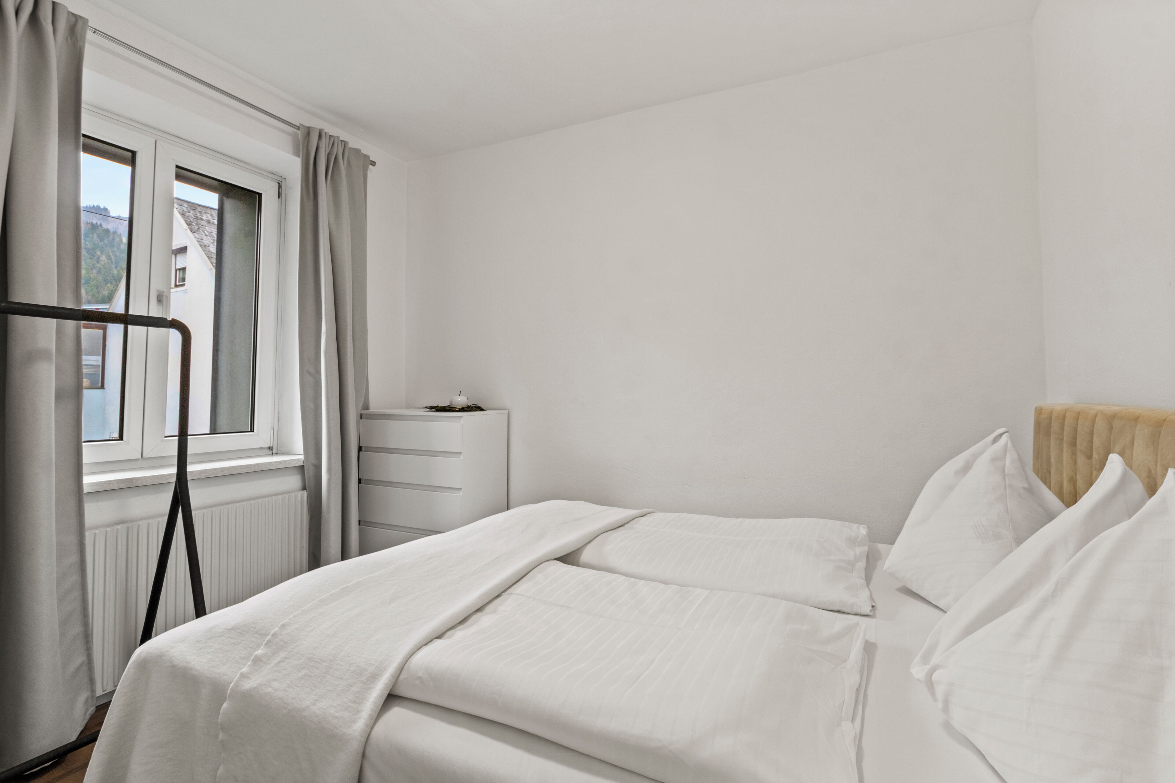 Ein Schlafzimmer mit einem Bett, einem Heizkörper, einem Fenster mit Vorhängen und einer kleinen Kommode.