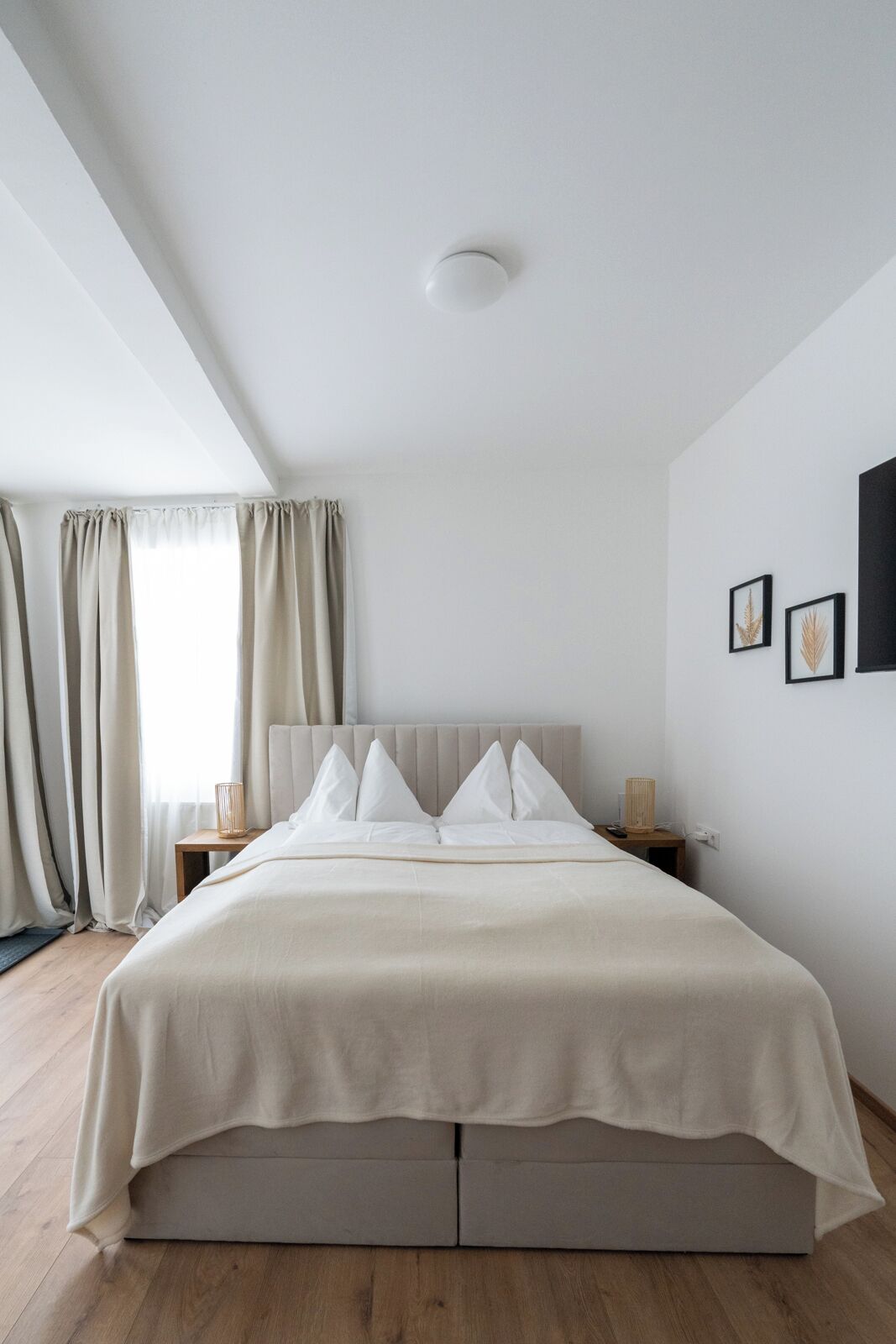 Ein schlicht eingerichtetes Schlafzimmer mit Bett, Vorhängen, Nachttisch und Wandbild.