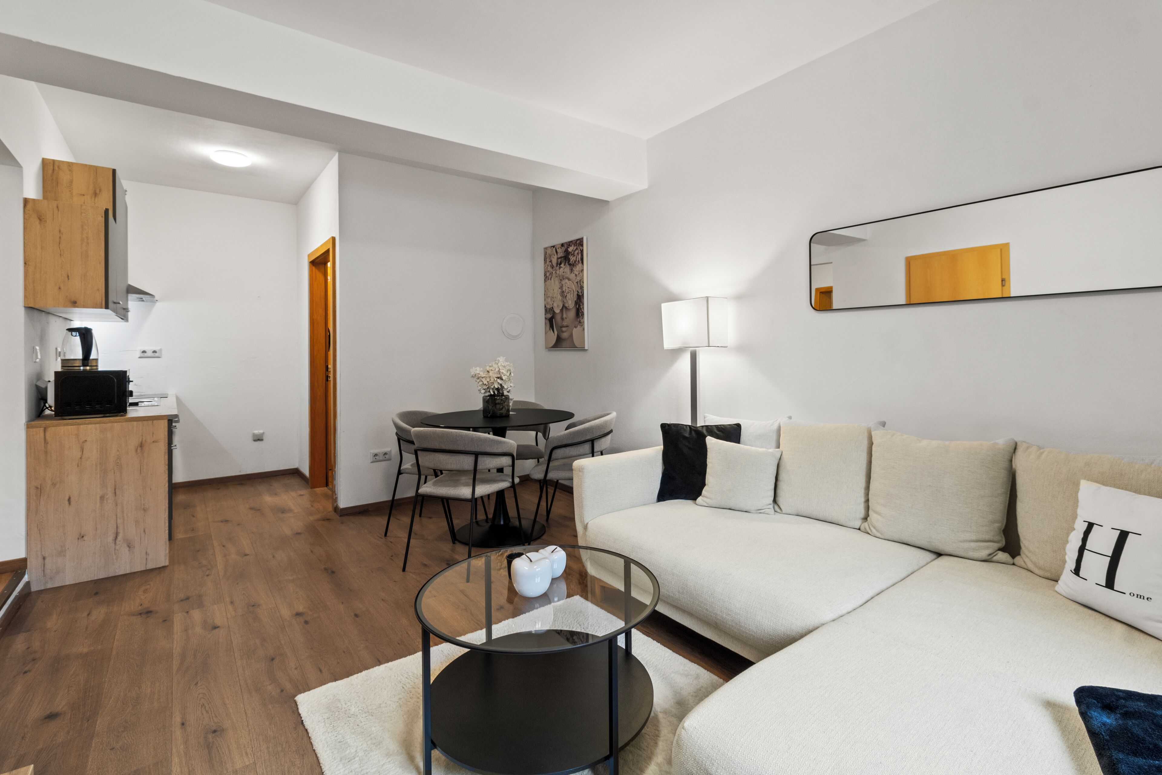 Ein modernes Wohnzimmer mit Sofa, Glastisch, Essbereich mit Tisch und Stühlen sowie Küchenzeile mit Schränken und Mikrowelle.