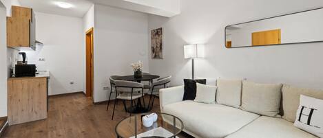 Ein modernes Wohnzimmer mit Sofa, Glastisch, Essbereich mit Tisch und Stühlen sowie Küchenzeile mit Schränken und Mikrowelle.