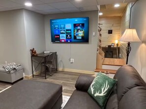 Ein Wohnzimmer mit flachbildfernseher an der Wand, einer Couch mit Zierkissen, einem kleinen Tisch mit Stühlen und einer Treppe im Hintergrund.
