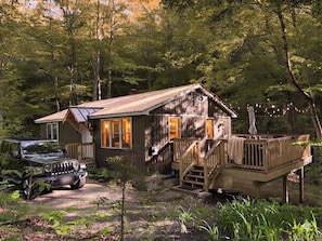 A rustic cabin with a wooden deck, a vehicle parked in front, and a forested backdrop.