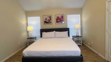 Une chambre à coucher avec un grand lit, deux tables de chevet, deux lampes fixées au mur et deux fenêtres équipées de stores.