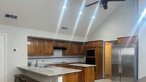 Una cocina con armarios de madera, un refrigerador de acero inoxidable, horno empotrado e isla de encimera de granito con taburetes de bar.