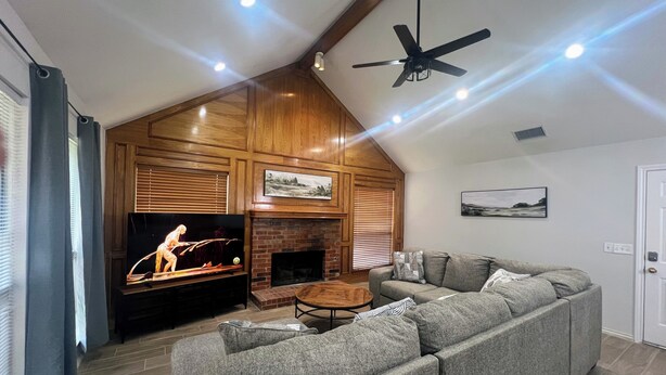 Una sala de estar con chimenea, televisor, ventilador de techo y un amplio sofá seccional gris.