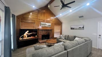 Un salon avec une cheminée, un téléviseur, un ventilateur de plafond et un grand canapé d’angle gris.