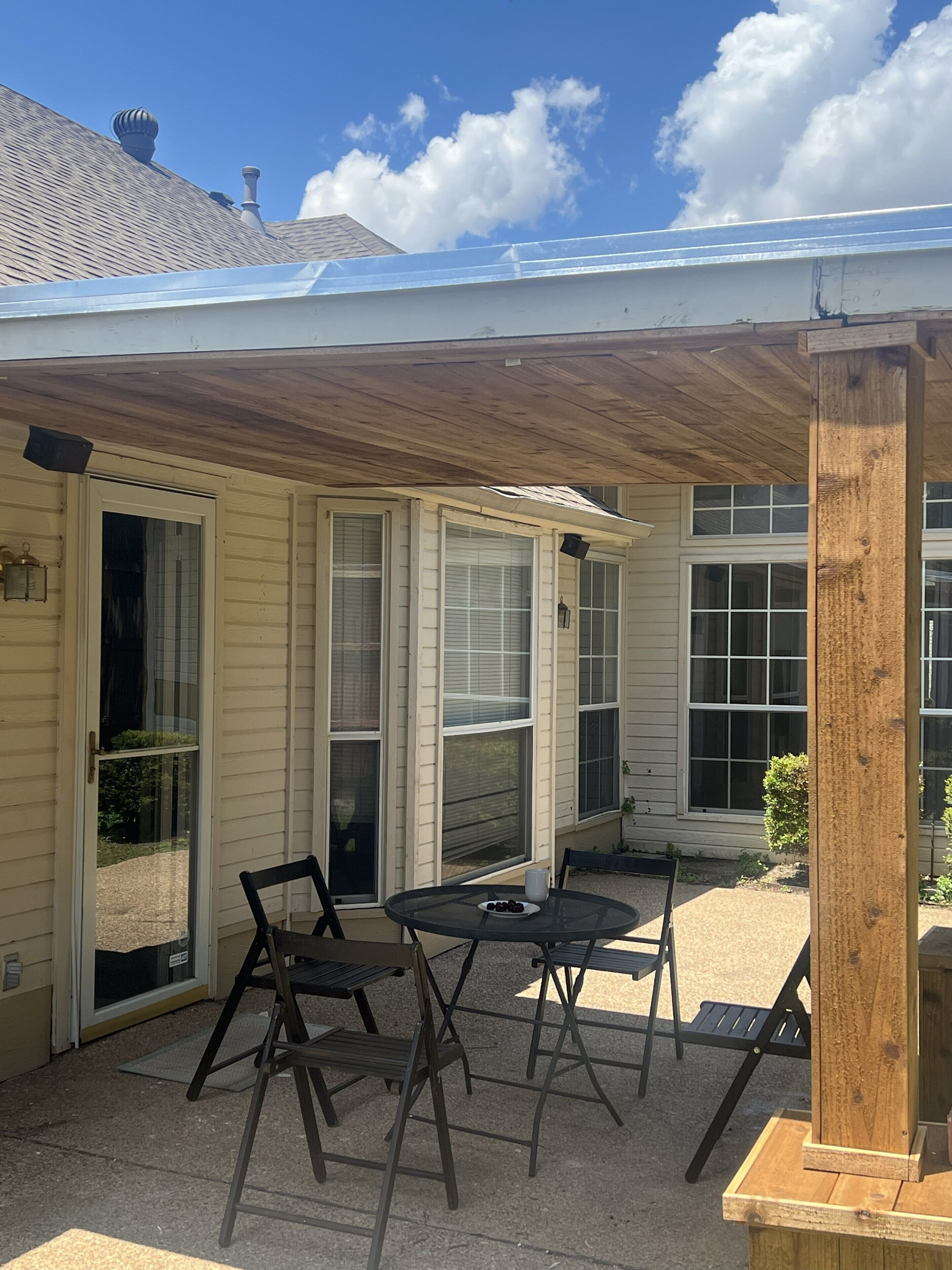 Une terrasse couverte avec une table et des chaises, un poteau en bois et une maison dotée de nombreuses fenêtres et d’une cheminée.