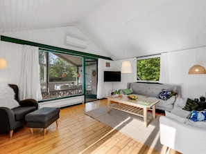 Ein modernes Wohnzimmer mit einer Couch, zwei Sesseln, einem Couchtisch und einem Fernseher. Ein großes Fenster bietet Blick auf Grünflächen.
