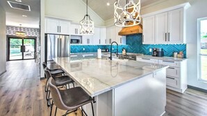 Une cuisine moderne dotée d’un grand îlot central, de tabourets de bar, d’appareils électroménagers en acier inoxydable et d’un éclairage décoratif.