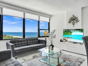 A modern living room with a sofa, coffee table, and TV, overlooking a beach. - Spacious Ocean View Condo / Beach Service / 704 (Miami Beach)