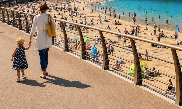 Une plage bondée, remplie de gens et de parasols, une femme et un enfant qui se promènent sur une promenade en bois, et une rambarde qui sépare la promenade de la plage.