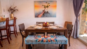 Een eetruimte met een houten tafel gedekt voor vier personen, een kleurrijke geweven bank en een schilderij van een boom op een klif.