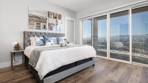 Ein Schlafzimmer mit einem großen Bett, einem Nachttisch, einem an der Wand befestigten Kunstwerk und einer Glastür, die auf einen Balkon mit Stadtblick führt.