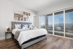 Ein Schlafzimmer mit einem großen Bett, einem Nachttisch, einem an der Wand befestigten Kunstwerk und einer Glastür, die auf einen Balkon mit Stadtblick führt.