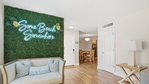 Una habitación con una pared verde, un letrero de neón que dice 'Some Beach Somewhere', suelo de madera, un sofá blanco con cojines estampados y una zona de comedor con candelabro.