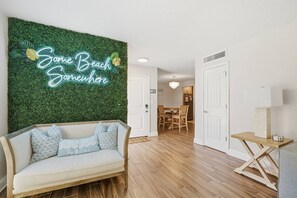 Ein Zimmer mit grüner Wand, einem leuchtenden Schild mit der Aufschrift 'Some Beach Somewhere', einem Holzboden, einem weißen Sofa mit gemusterten Kissen und einem Essbereich mit einer Chandelier.