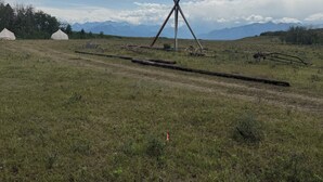 Um campo gramado com uma estrutura de moinho de vento, tendas e montanhas ao fundo.