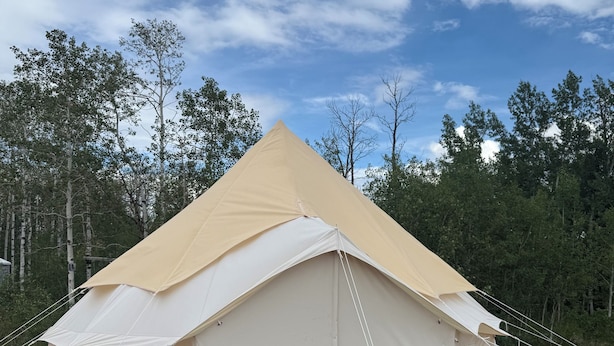 Uma tenda grande, bege e em formato de cúpula, com base branca, colocada em uma plataforma de madeira com grama sintética verde, cercada por árvores.