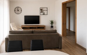 Ein Wohnzimmer mit einer Couch, Sesseln, einem Fernseher, einer Uhr und einer Pflanze.