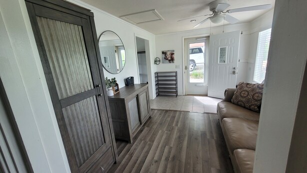 A hallway with a wooden floor, a ceiling fan, a door with a mirror, a small shelf with plants, and a couch with patterned cushions. - Kansas City 3-bedroom farmhouse on peaceful acreage near Legends entertainment! (Kansas City)