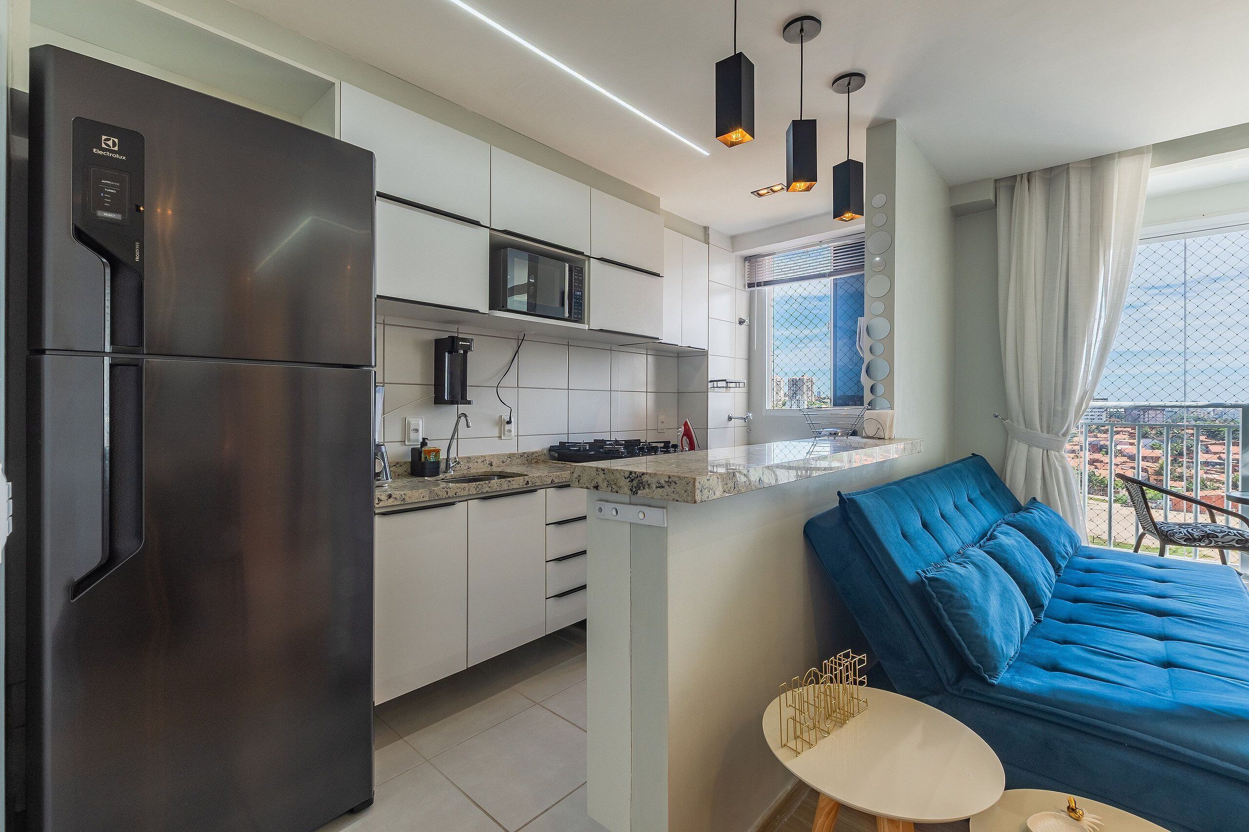 A modern kitchen with a built-in microwave, stainless steel refrigerator, and a dishwasher. There is a dining area with a blue sofa and a small table.