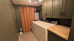 A compact laundry room with a washing machine, wooden cabinets, and a ceiling light fixture. - The Green Clover Hideaway-Cozy Studio Retreat with Fire Pit (Draper)