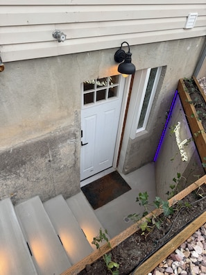 Exterior de formigó amb una porta blanca, una finestra petita i una jardiniera de fusta amb vegetació.
