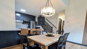 A dining area with a wooden table and chairs, a chandelier, and a kitchen in the background. - Three Peaks Penthouse [3 Hot Tubs & Pool] (Canmore)