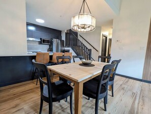 Ein Essbereich mit Holztisch und -stühlen, einer Chandelierleuchtre und einer Küche im Hintergrund.
