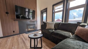 A modern living room with a fireplace, a sofa, a small round table, and a television mounted on the wall. - Three Peaks Penthouse [3 Hot Tubs & Pool] (Canmore)