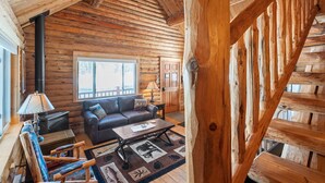 Interno di una baita di legno con camino, divano, tavolino e vista sulla natura circostante.