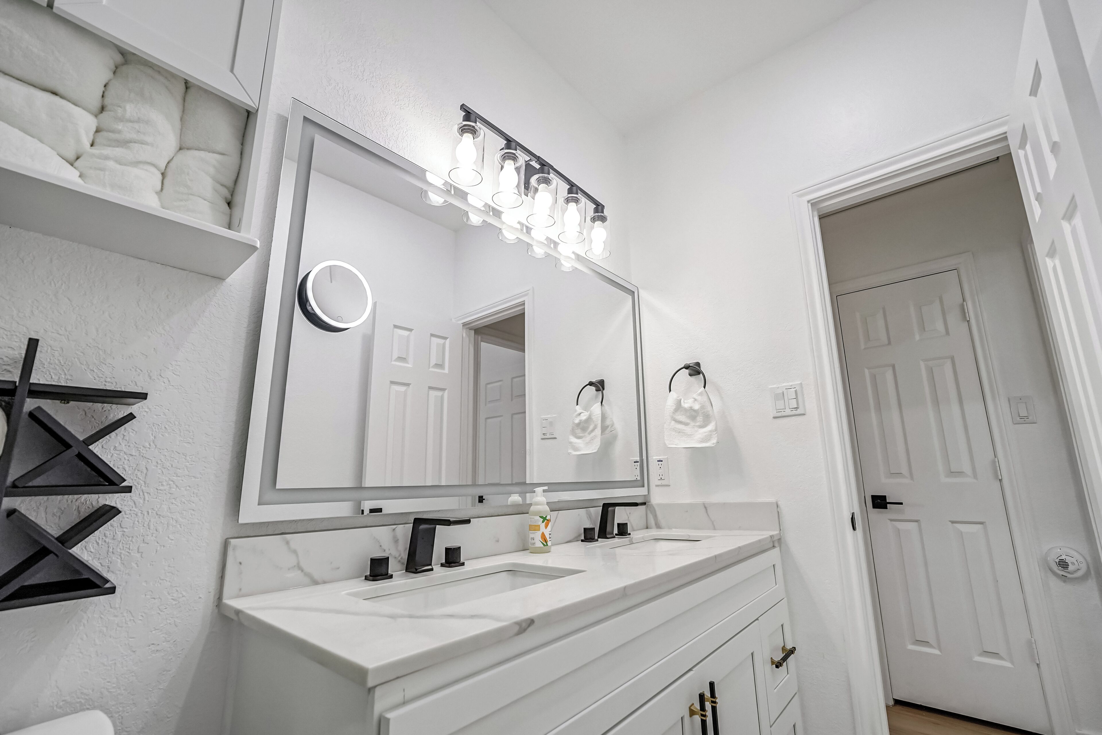 Un baño con un espejo grande, dos lavamanos, un perchero para toallas y una puerta que da a otra habitación.