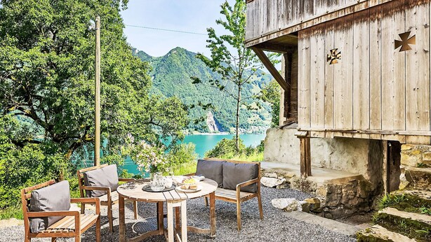 Eine Holzterrasse mit Tisch und Stühlen mit Blick auf einen See und bewaldete Berge.