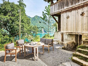 Um deck de madeira com mesa e cadeiras, com vista para um lago e montanhas arborizadas.