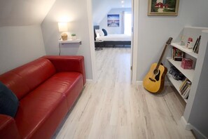 Una habitación con sofá rojo, una guitarra, una estantería y una cama con vista a una casa a través de un puerta abierta.