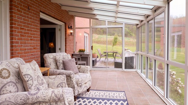 A covered patio with a brick wall, a sofa with patterned cushions, and a view of a garden. - The Homestead at Anderson Ridge |Pool & Trails (Schellsburg)