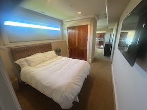 Ein Schlafzimmer mit einem Bett, einem Nachttisch, einer Lampe, einer Holztür und einem Flur, der in eine Küche führt.