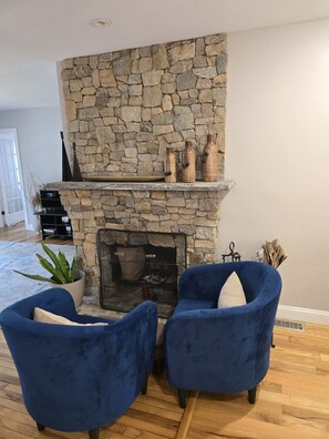 A stone fireplace with a mantel, two blue armchairs, a potted plant, and a bookshelf.
