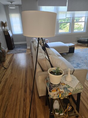 A living room with a sofa, a floor lamp, a coffee table with a plant, and a view of the outdoors through large windows.