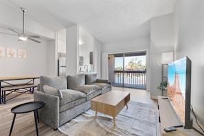 A modern living room with a grey sofa, a wooden coffee table, a black side table, a ceiling fan, a flat-screen TV, and a sliding glass door leading to an outdoor area.