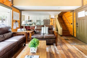 A living room with a brown leather sofa, a wooden coffee table with a plant, and a kitchen in the background. - Quiet Cul-De-Sac, Walk to SHARC w/ free passes, Hot Tub, AC, Sleeps 13! (Sunriver)