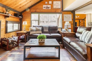 A cozy cabin interior with a wooden ceiling, a large window, a brown leather sofa, a coffee table, and a fireplace. - Quiet Cul-De-Sac, Walk to SHARC w/ free passes, Hot Tub, AC, Sleeps 13! (Sunriver)