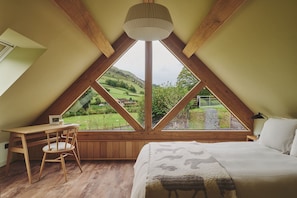Ein Schlafzimmer mit großem Fenster, Holzbalken, einem Bett, einem Sessel und einem Schreibtisch.