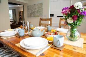 A breakfast table set with plates, mugs, a teapot, a coffee pot, a fruit bowl, and a vase of flowers. - Grimsbury Castle - Luxury Cottages (Hermitage)